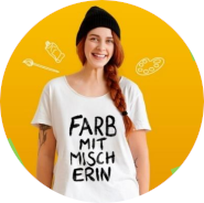Frau mit rotem langen Zopf steht lächelnd vor einem orangen hintergrund. Sie hat ein weißes T-Shirt an mit der Aufschrift "Farbmitmischerin" und eine schwarze Wollmütze auf dem Kopf.